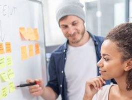 Two adult students collaborate in a modern workspace, discussing ideas on a whiteboard covered with colorful sticky notes.