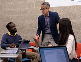 Male professor and 2 students engage in a discussion around a table, with laptops open and notes visible in a collaborative setting.