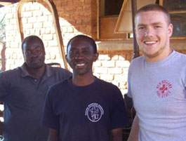 3 individuals stand together outdoors in front of a brick building, dressed casually and smiling for the photo.