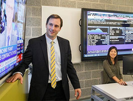 A business professor stands next to a large screen displaying financial news, while a female student sits at a desk with a computer.