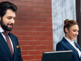 Two hotel receptionists in professional attire stand behind a front desk, surrounded by a modern brick wall and ambient decor.