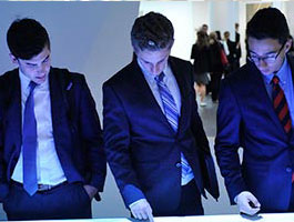 3 male students in suits engage with a digital display, surrounded by a lively, modern atmosphere in a professional setting.