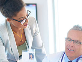A professional woman in a gray blazer engages in a discussion with a male doctor in a bright medical office.