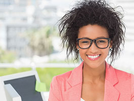 Female student with curly hair wearing a coral blazer sits in an office setting with a blurred cityscape in the background.