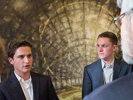 2 students in suits engage in conversation in front of an abstract, textured background.