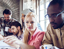 A group of five adult students engaged in a collaborative discussion around a table, with coffee cups and notebooks scattered about.