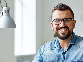 A male adult learning student sits at a desk with a modern lamp in a well-lit room, wearing a denim shirt, focused on his work.