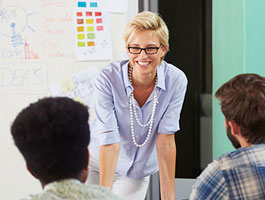Female professor engages with students in a classroom setting, in front of a whiteboard filled with notes and colorful visuals.