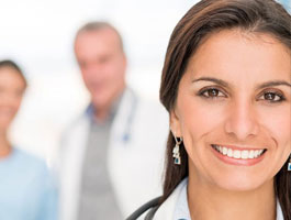 A woman with dark hair and a stethoscope around her neck stands in a medical setting, alongside two healthcare professionals in the background.