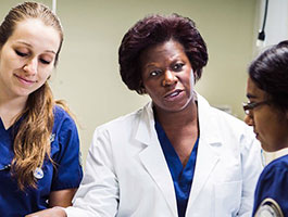 1 professor and 2 nursing students in blue scrubs and a white coat collaborating in a clinical setting.