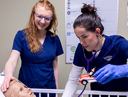 2 nursing students in scrubs are tending to a medical simulation patient, one using a stethoscope on a toy car.