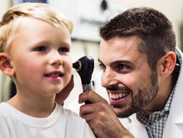 A doctor examines a child's ear with an otoscope in a clinical setting. The child wears a white shirt and looks attentive.