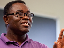A male professor in a purple shirt gestures while speaking, wearing glasses, in a bright, modern indoor setting.
