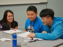 Female teacher and 2 students gathered around a table, engaging in a group study session with books and notes spread out.