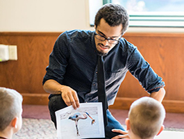 Teaching student in a dark shirt kneels, showing a colorful bird illustration to a group of attentive children sitting on the floor.