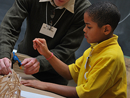 A male teacher in a green sweater assists a child in a yellow shirt with a hands-on learning activity, using tools and materials.