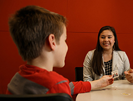 A young boy in a red and gray shirt reaches out during a seated conversation with a female teacher at a table against a red wall.